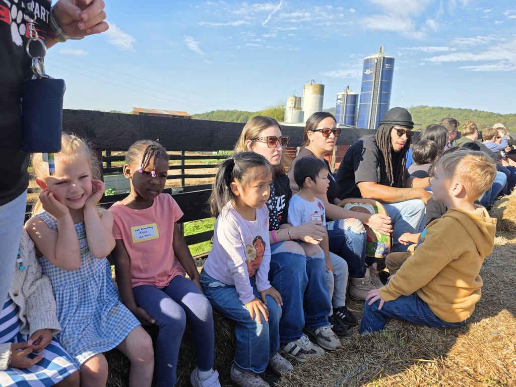 A group of children and adults sit on hay bales at an outdoor event. The sky is blue.