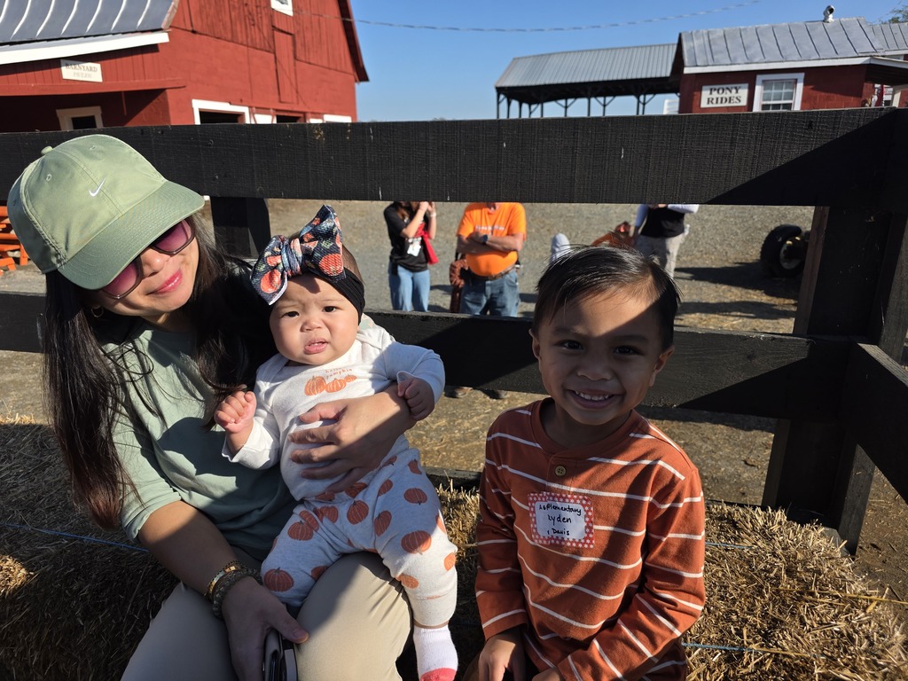 A woman in a hat holds a baby while a child stands next to them, all smiling in a hay field.