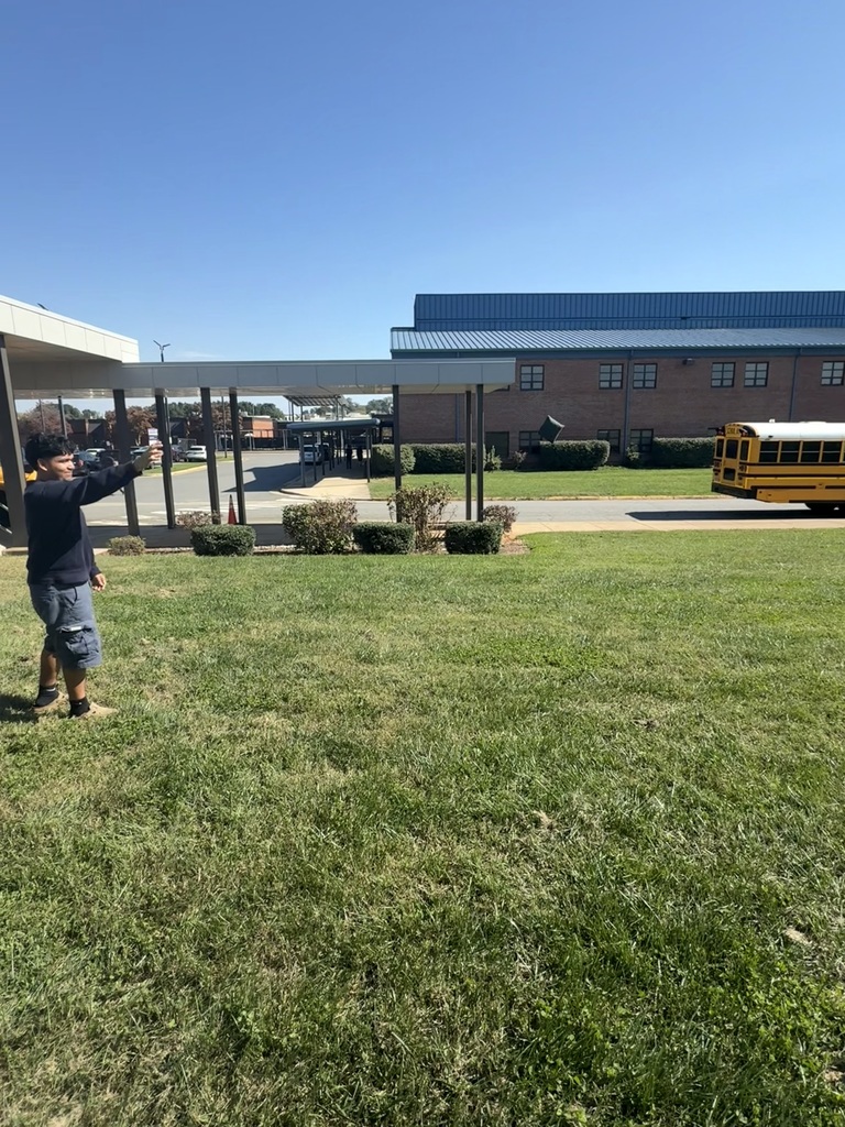 A person stands in a grassy area in front of a school, with a yellow bus in the background.