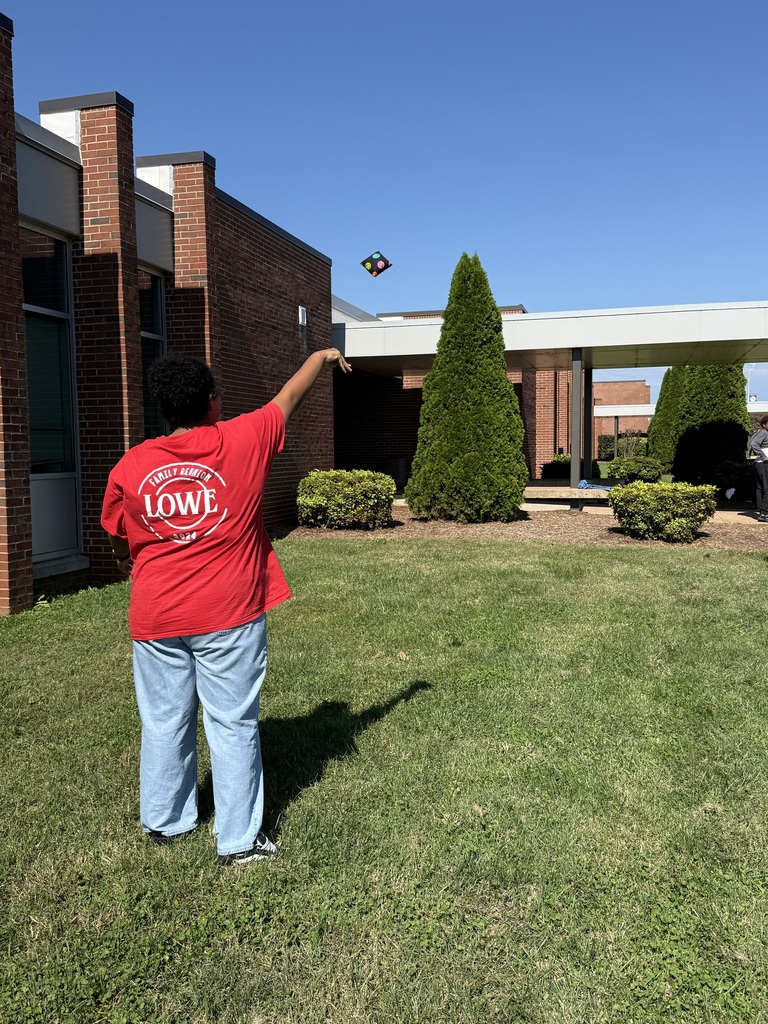 Person in red shirt and blue pants on a lawn, pointing to the sky. Buildings and greenery in the background.