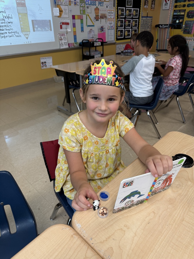 A young girl in a yellow dress wearing a crown sits at a desk with a book.