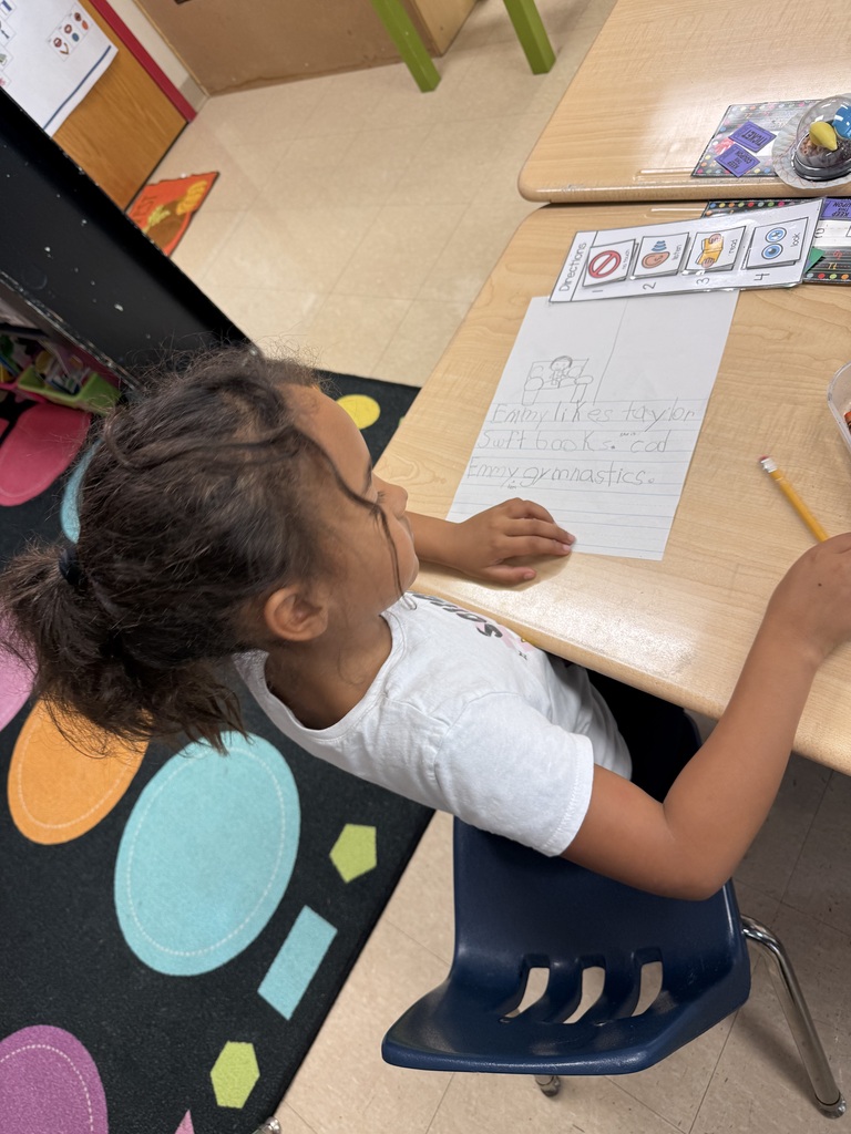 A young child, sitting at a desk, is focused on writing on a paper while holding a pencil.