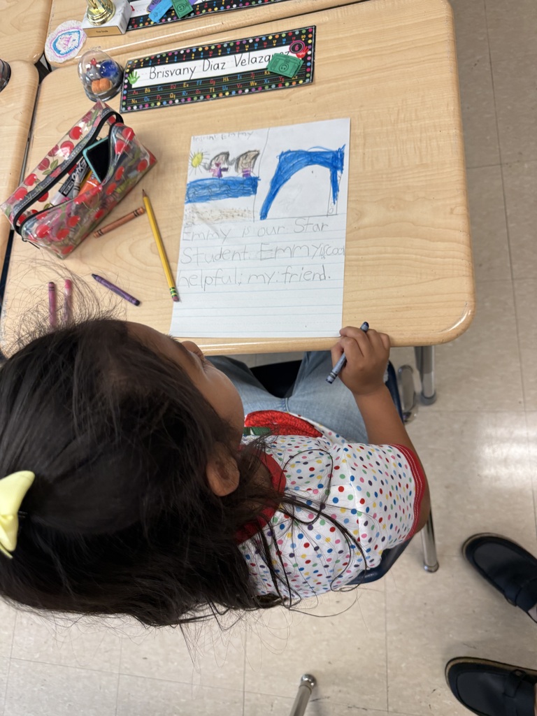 A child writes on a paper at a desk with pencils, a pouch, and a book.