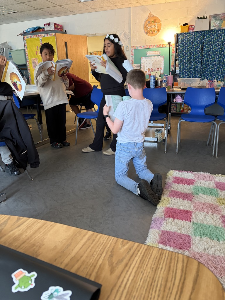 A classroom with a child kneeling and others standing. They hold books and read. Blue chairs and desks are present.