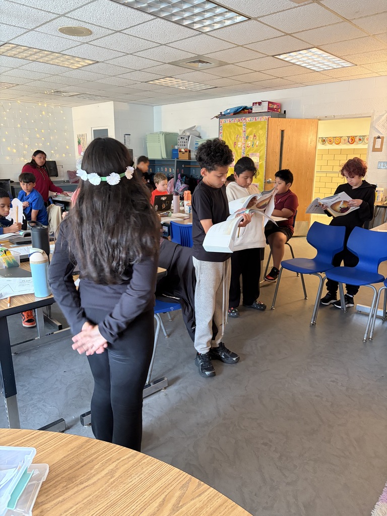 A classroom with students reading books, a woman standing, and others sitting on blue chairs.