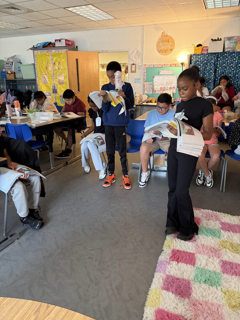 Several children in a classroom sit at desks or on chairs, reading books. One child stands with a book.