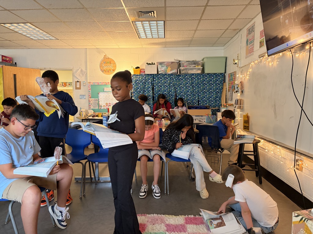 A group of students in a classroom. Some are seated while others stand, holding books. A student stands in front, likely leading a discussion.