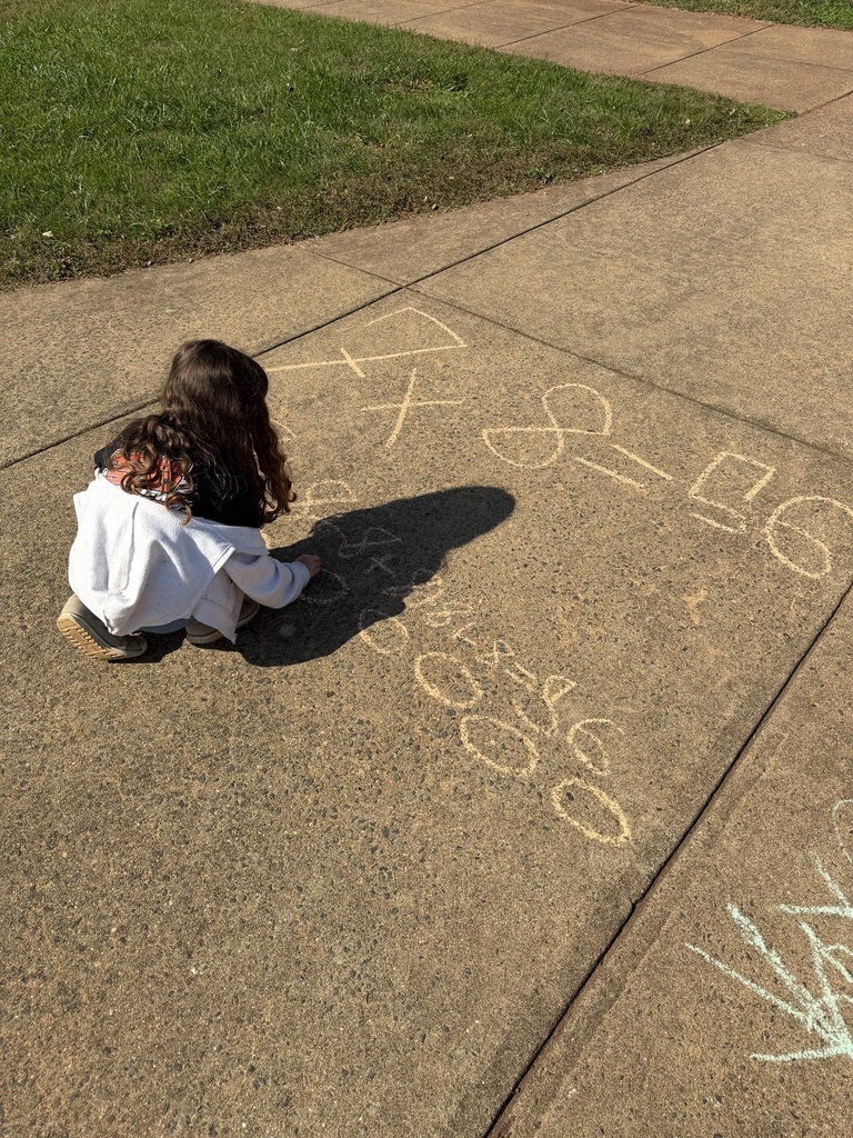 A child is kneeling on the sidewalk, drawing numbers and letters in chalk. Background is a grassy area.