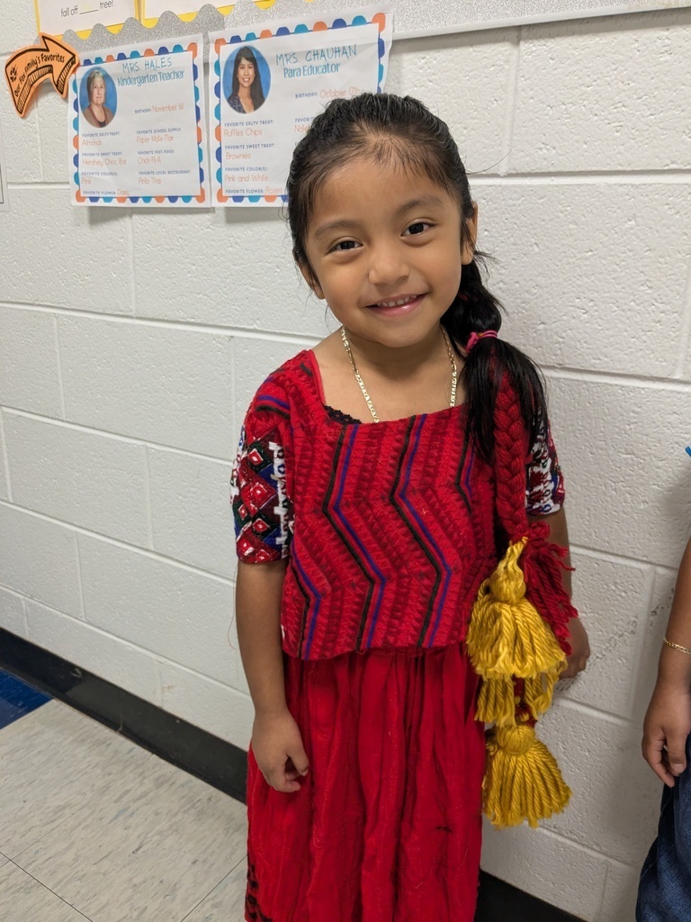 A young girl in a red dress with yellow tassels smiles in a room with white walls and bulletin boards.