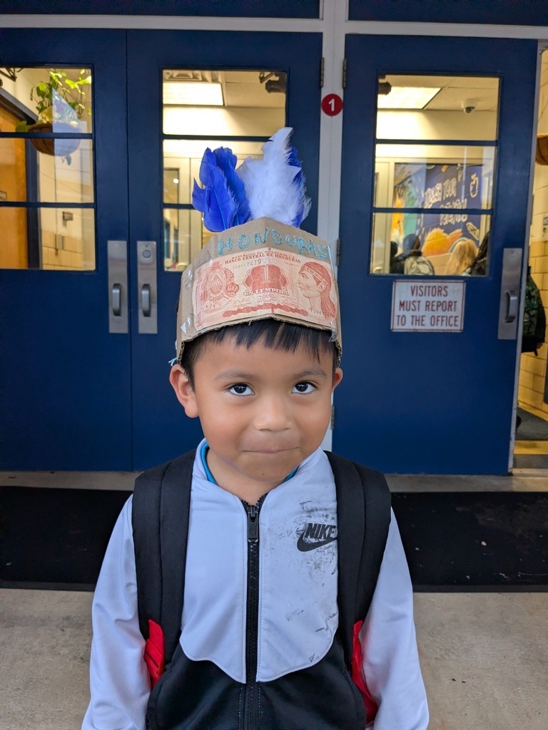 A child stands in front of a blue double door, wearing a backpack and a colorful hat with feathers.