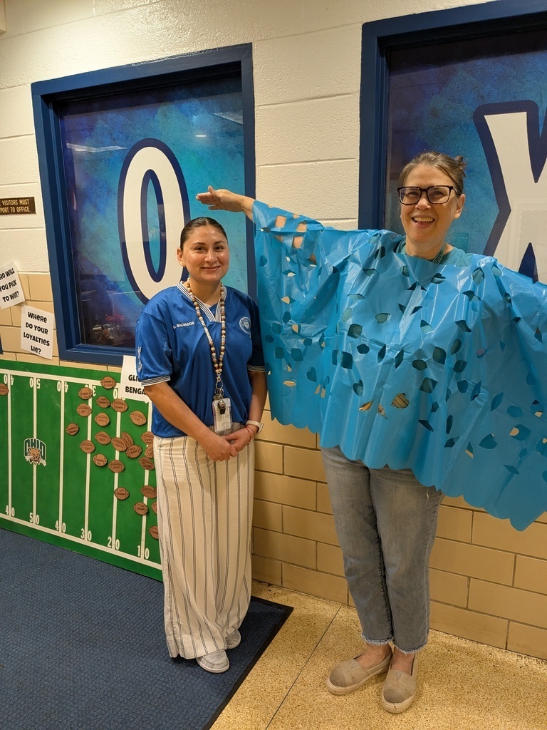 Two women in a corridor, one wearing a blue cape with cutouts, standing near a green wall with coins.