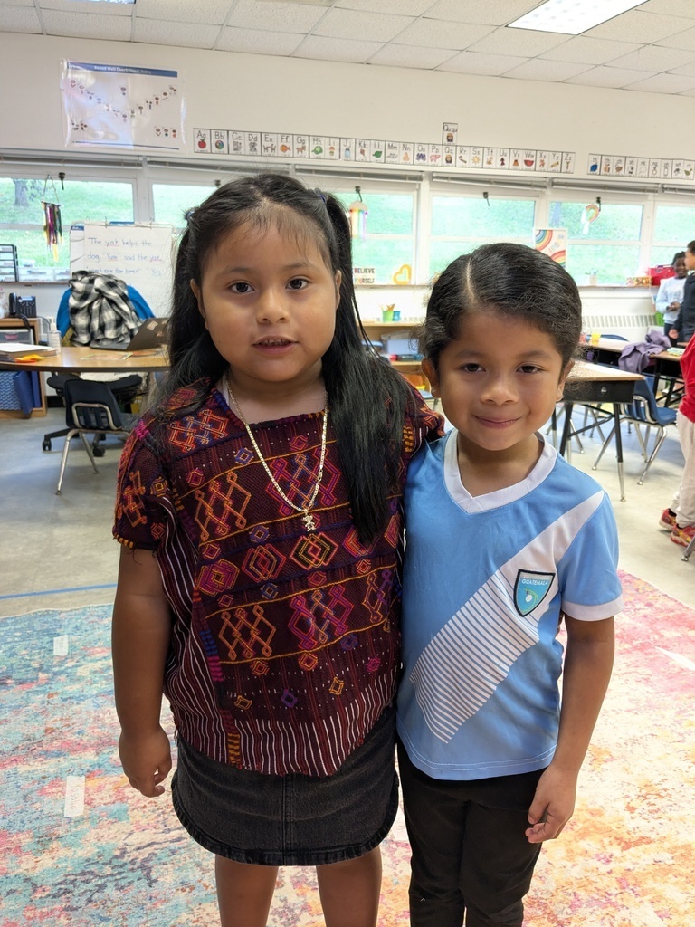 Two children stand in a classroom, a girl with dark hair and a boy in blue. Behind them are tables and chairs.