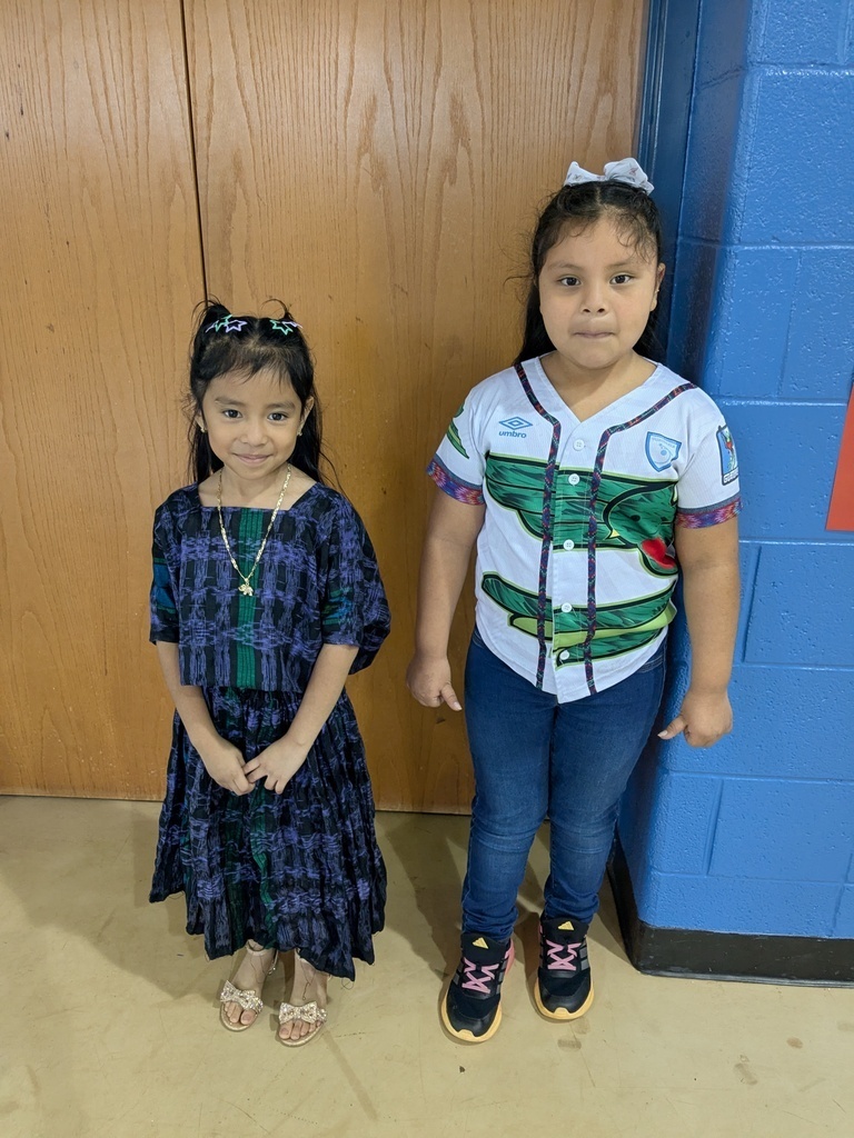 Two young girls stand in a room. One wears a blue dress and sandals, the other a white jersey shirt and sneakers. Behind them, a blue wall meets a wooden door.