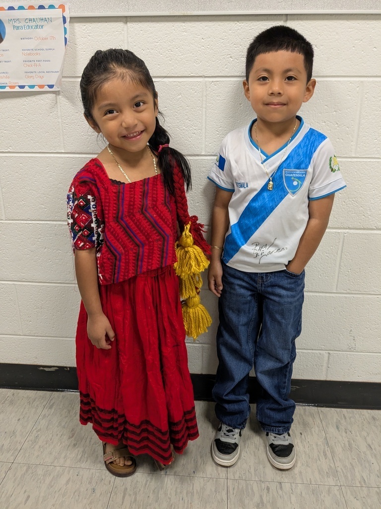 Two children, a girl in a red dress and a boy in a white and blue shirt, stand next to each other.
