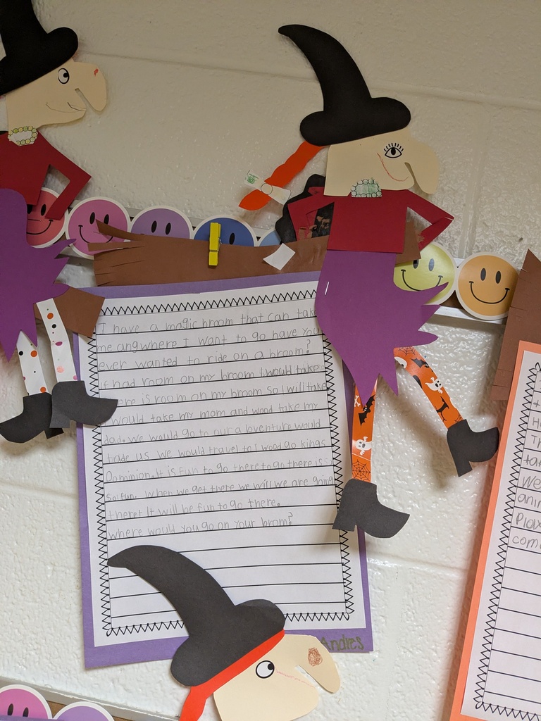 Paper cutouts of witches and smiley faces on a white wall. One cutout is holding a paper with writing.