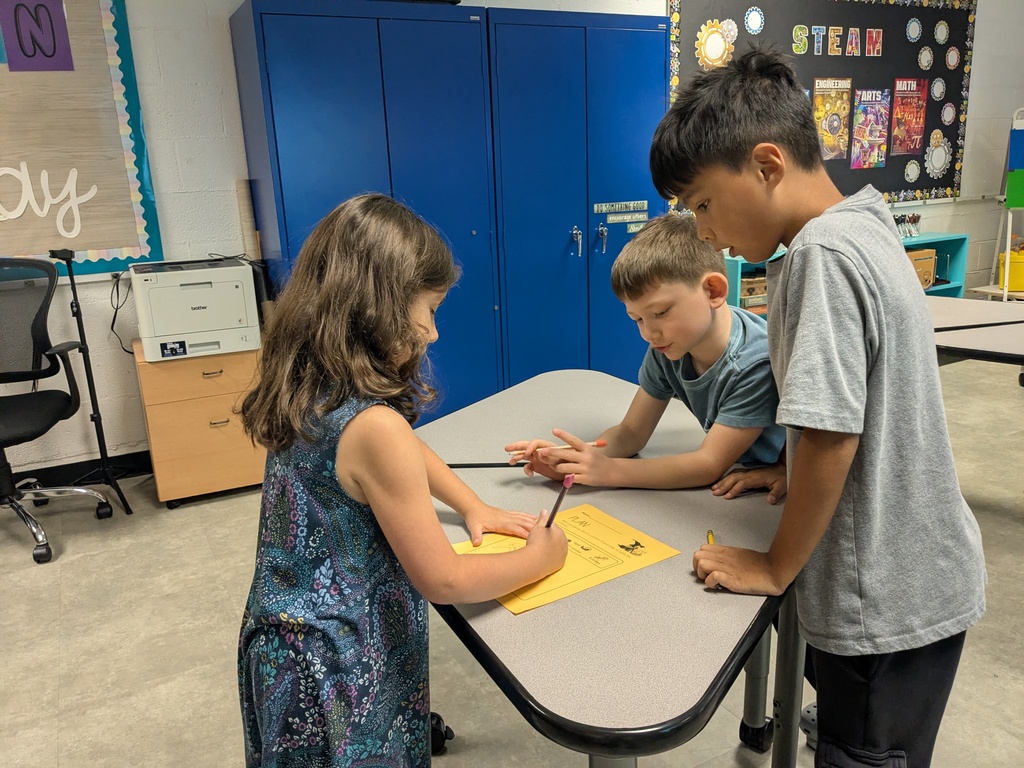 Three children in a classroom, focused on a yellow paper and writing utensils on a table.