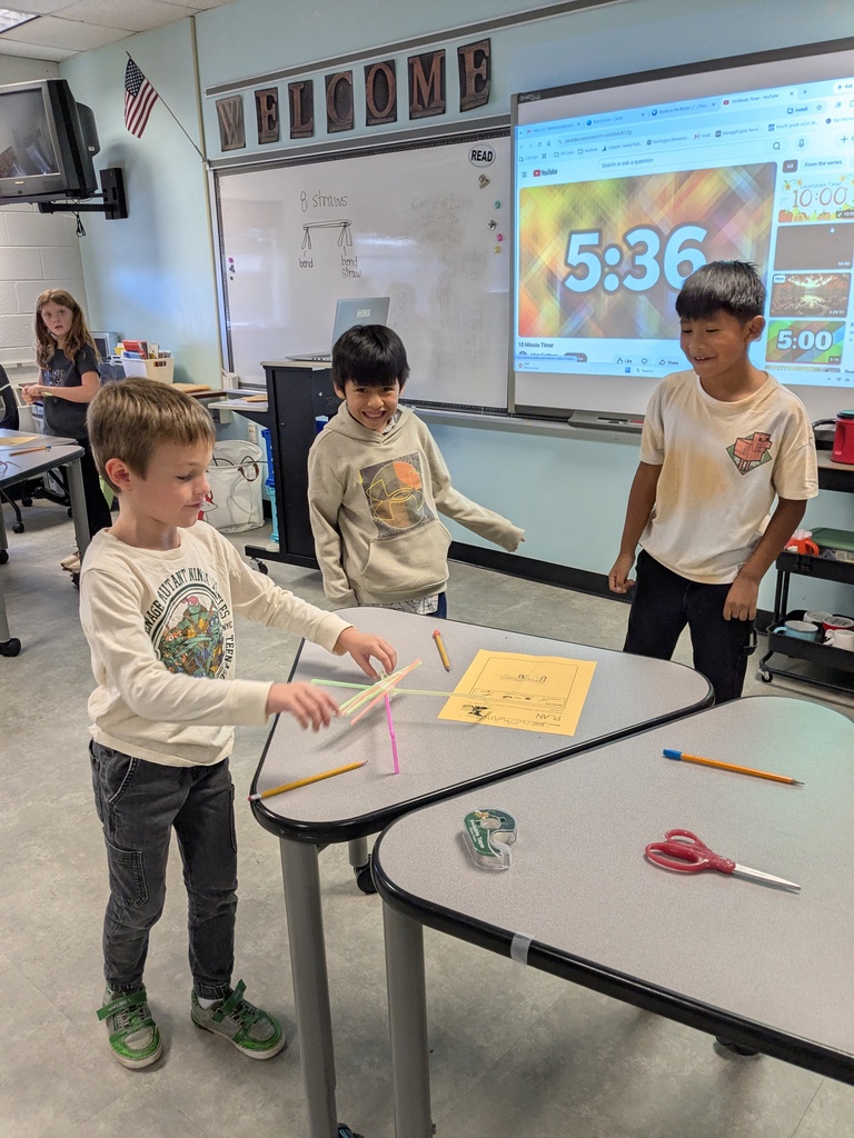 Three children at a desk with a yellow paper and craft materials. Behind them, a whiteboard with the time 5:36.
