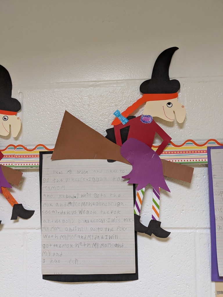 A classroom wall displays a paper cutout of a witch, in black and purple, holding a broom. The witch wears a hat and a ribbon.