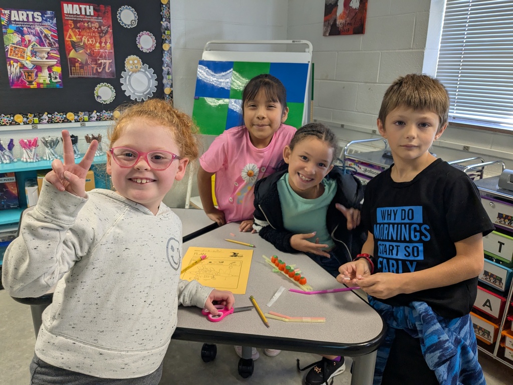 A classroom with kids smiling. One child is holding scissors. Behind them is a table with art materials.