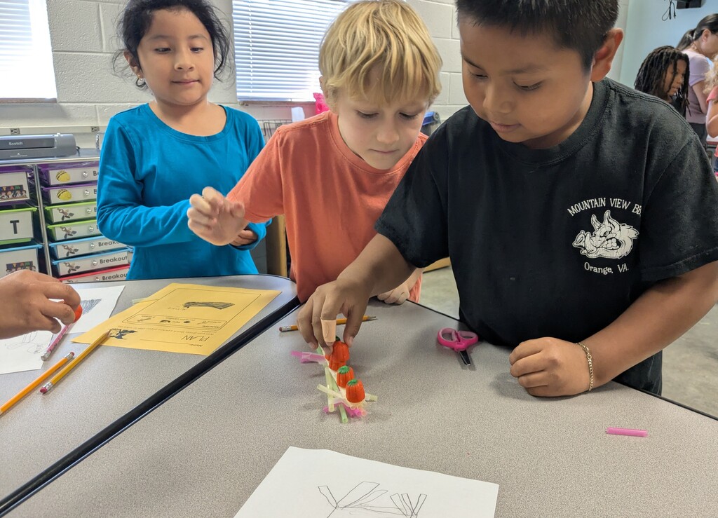 Children are gathered around a table in a classroom. They are engaged in an activity involving colorful objects.