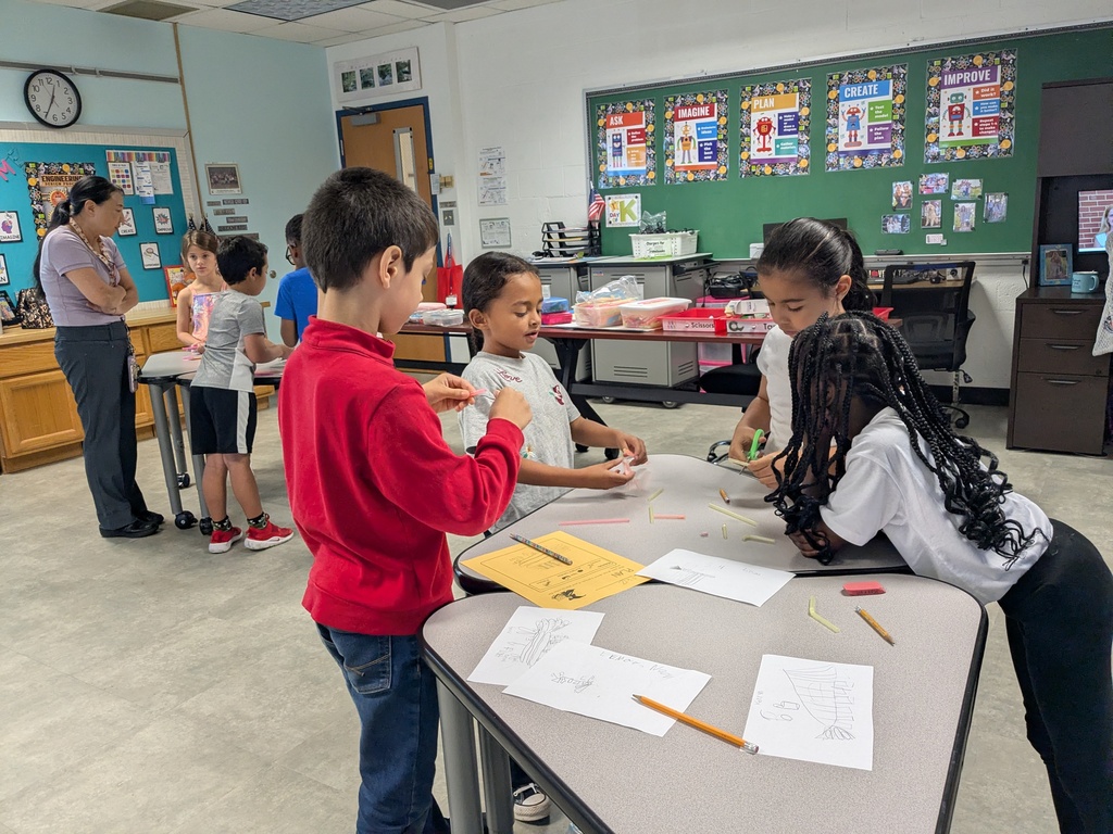 Kids work together at a table in a classroom. Papers, pencils, and a clock on the wall.