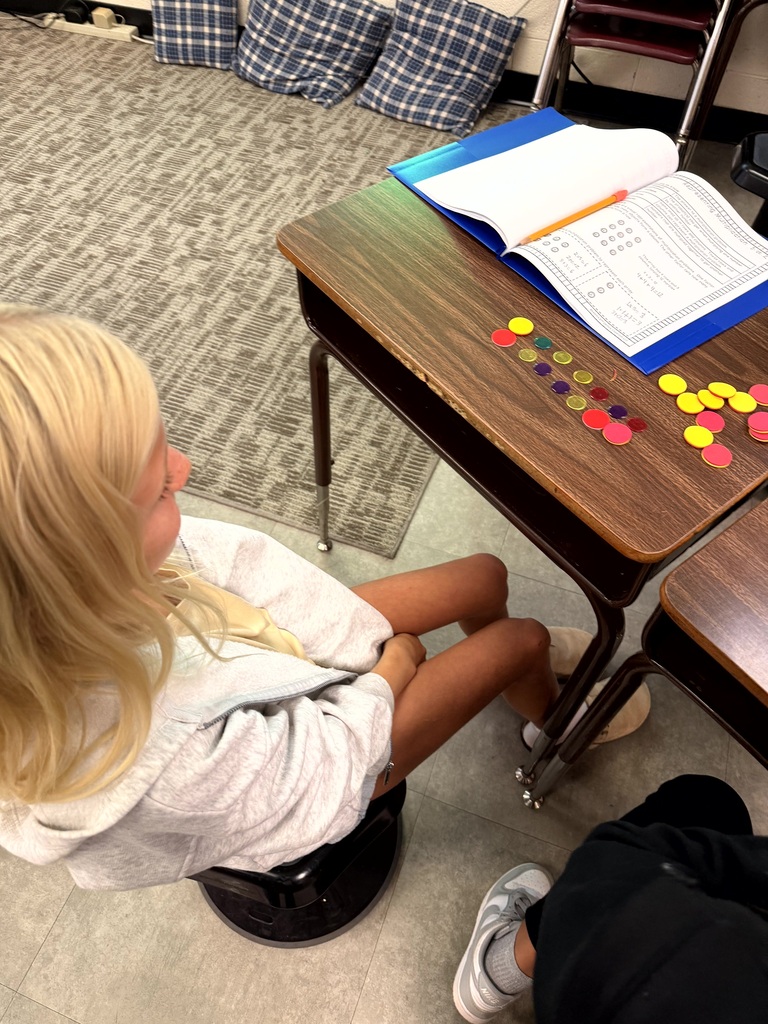 A student with blonde hair sits cross-legged on a chair in front of a desk.