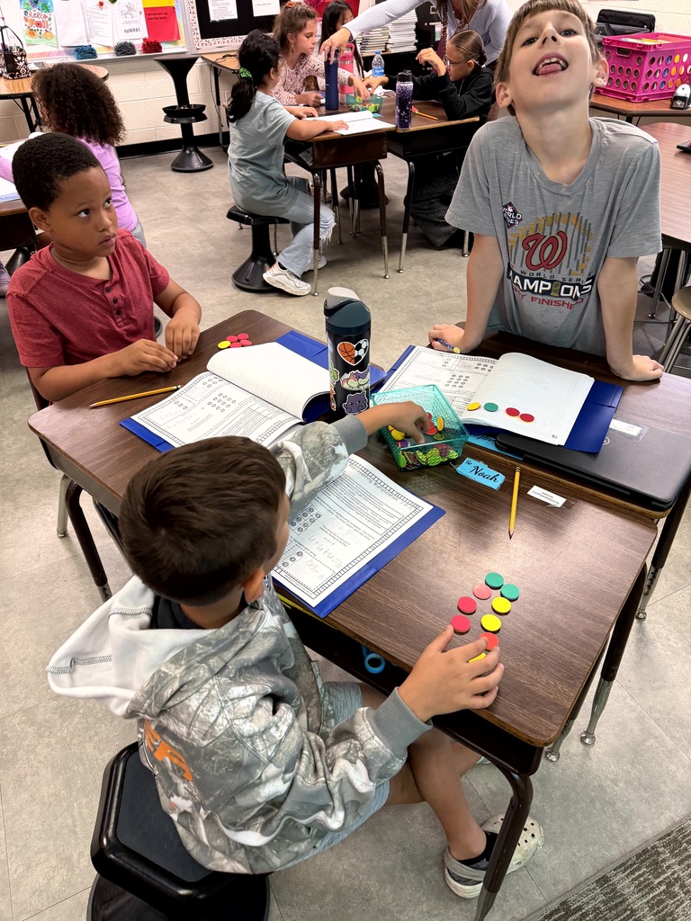 Four children work on schoolwork at desks in a classroom with a white and blue wall.