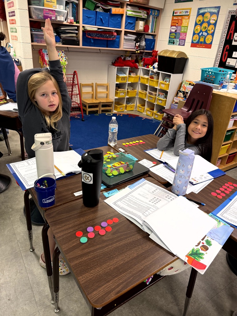 Two children sit at desks in a classroom. One child has her hand raised. Papers, pens, and other items are on the desks.