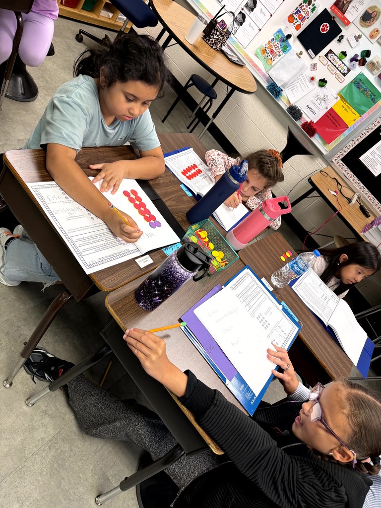 Several students work on papers at desks with an adult. Tables have binders, books, and water bottles.