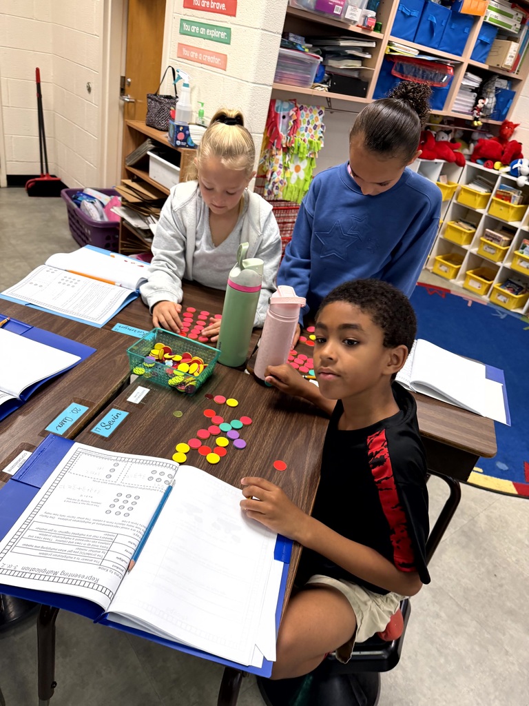 Three children work at a table with books, papers, and colorful objects, with shelves in the background.