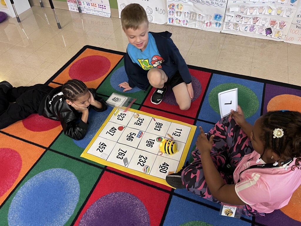 Three children playing a game on a colorful floor mat, with one holding a paper.