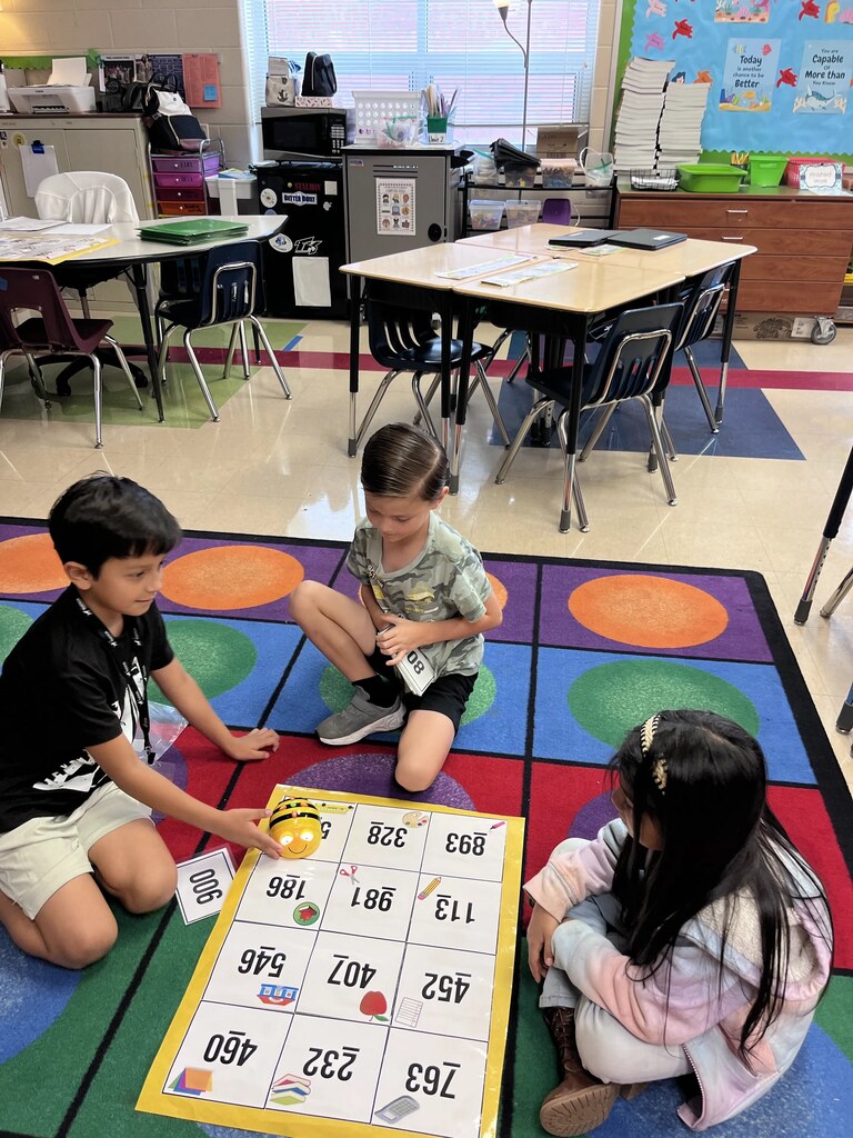 Three children sit on a floor, playing a game. One holds a board with colorful letters and numbers.