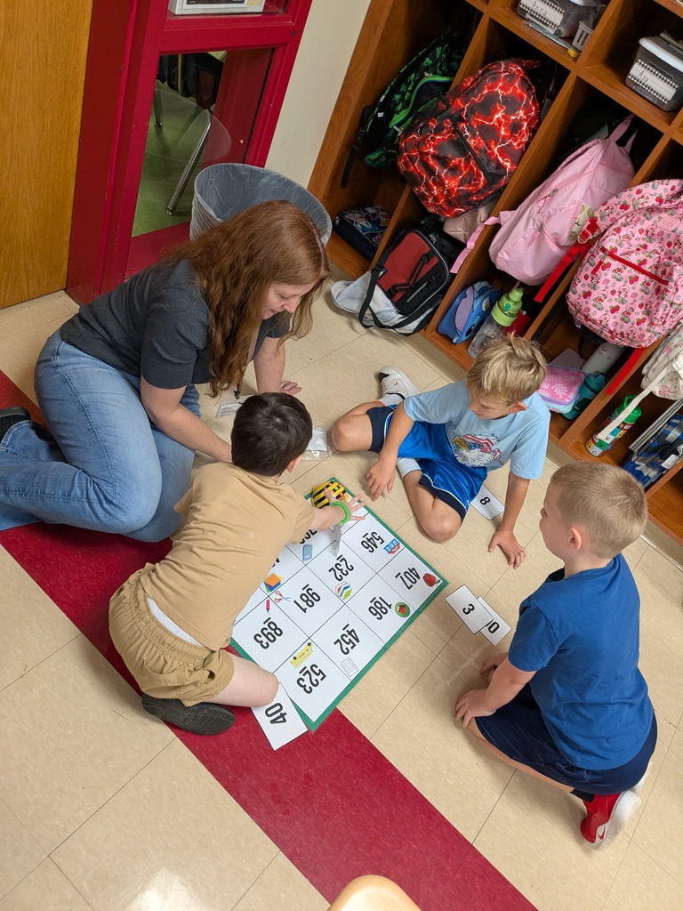 A woman and three children are seated on the floor, interacting with a number grid. They are in a room with shelves of bags and other items.