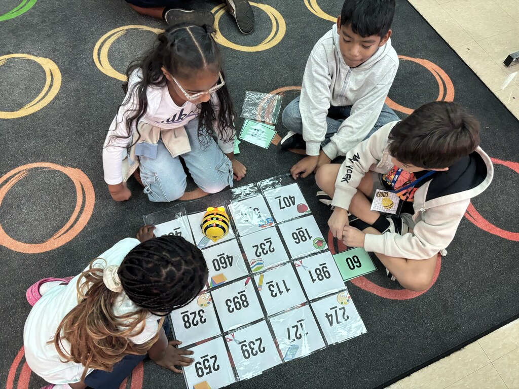Four children, wearing jackets, play a game with numbers on the floor. A yellow toy is nearby.