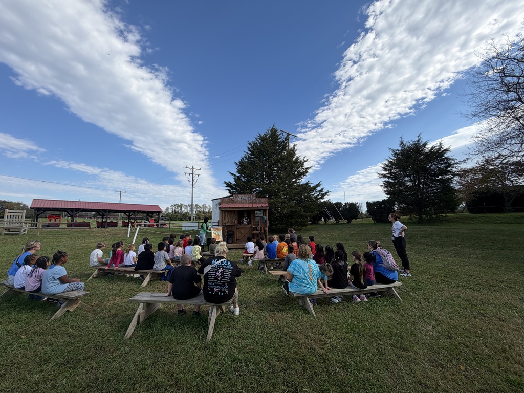 A group gathers on benches in a grassy field under a blue sky. A person stands before them.