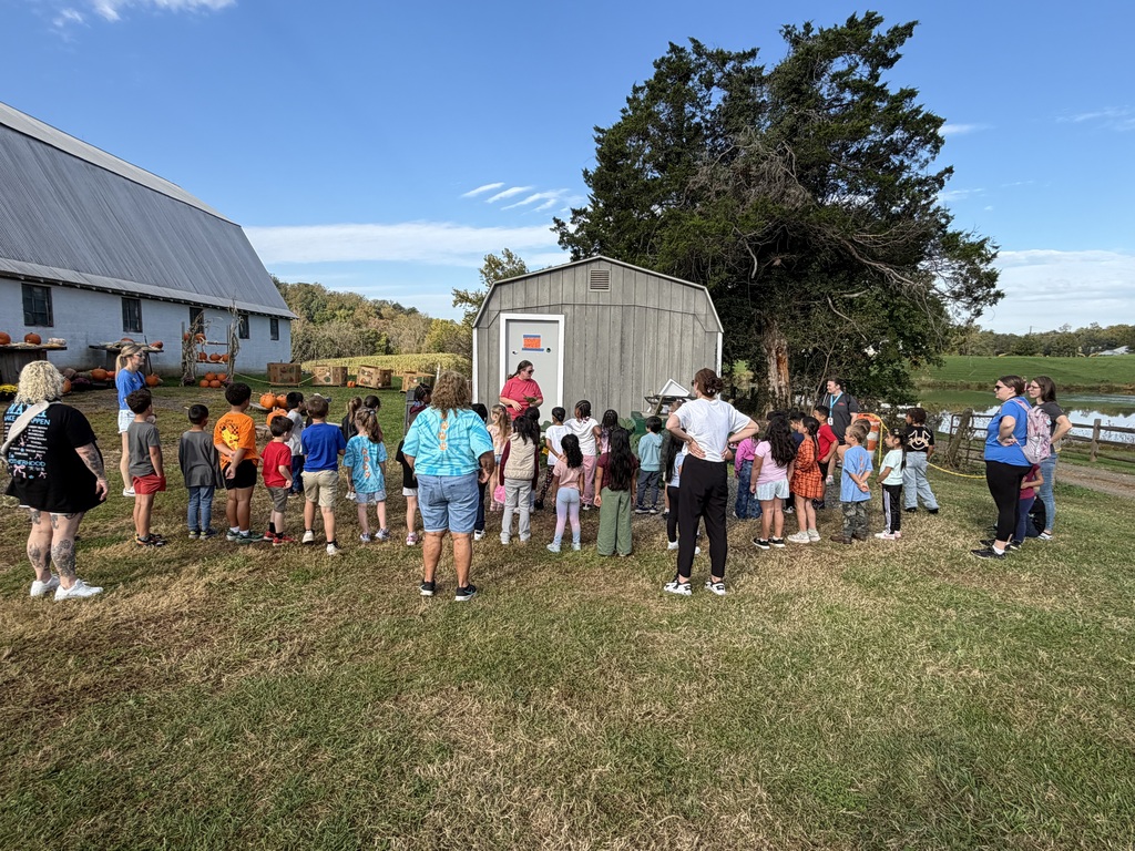 Group of people, some in school uniforms, standing in a field with a barn and trees in background.