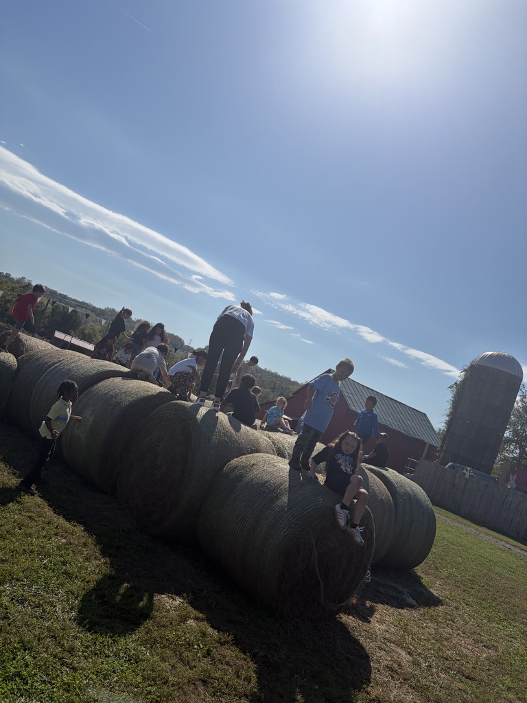 Several people are on top of stacked hay bales on a farm with a silo in the distance.