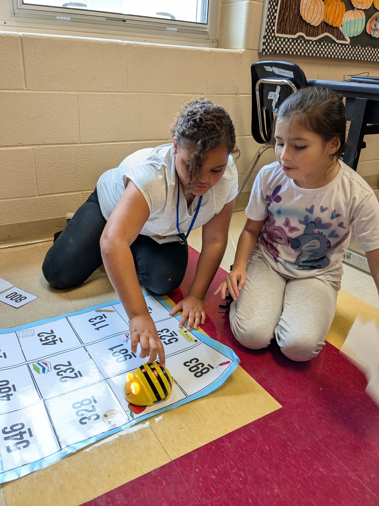 Two children playing with a robot on a mat with numbers. One is crouched down while the other sits.
