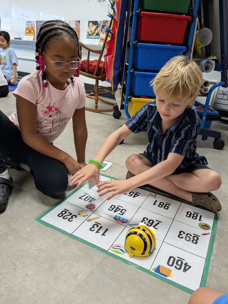 Two students work on a number grid with a toy on the ground, seated on floor.