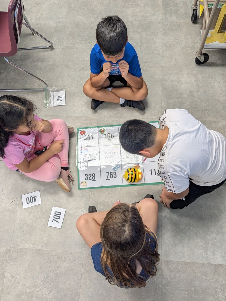 Four children sit on the floor around a game board with numbers and a bee.