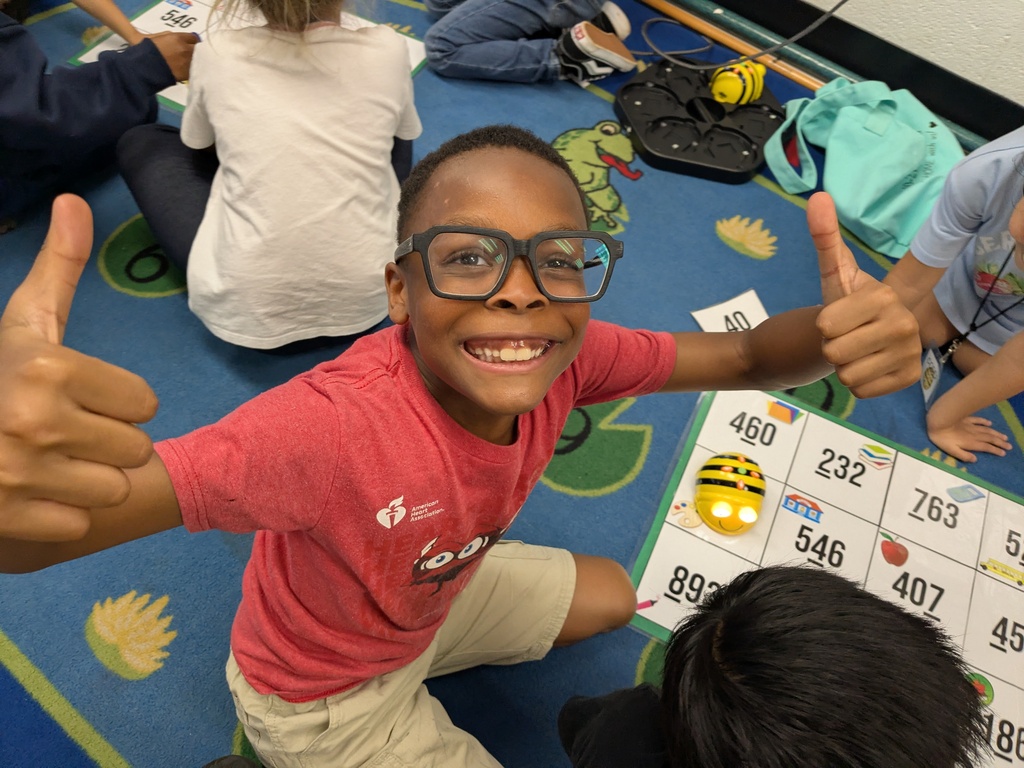 A boy in a red shirt gives two thumbs up while kneeling next to a board with numbers.
