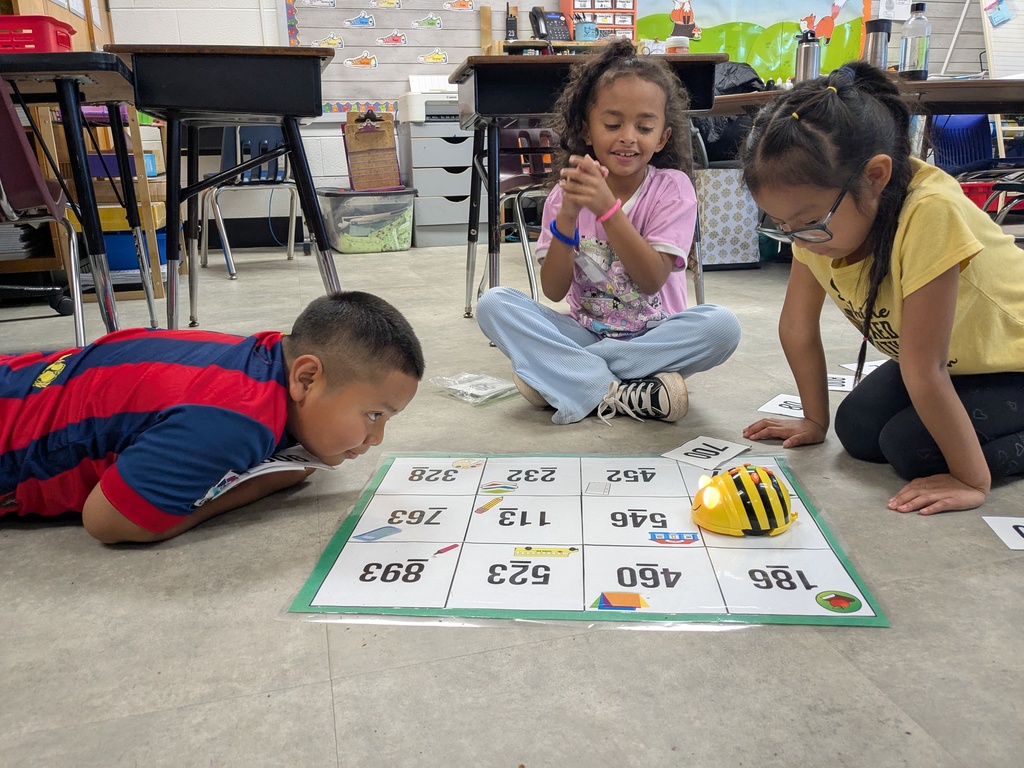 Three young children playing a board game with number tiles on the floor in a classroom.