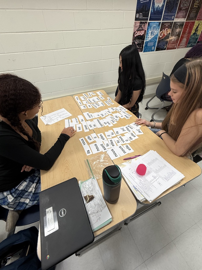 Three students seated around a table, with papers and cards spread out, discussing while using a laptop.