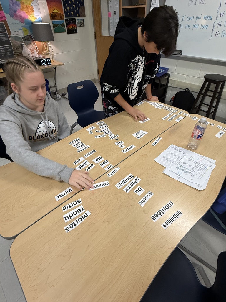 Two students work at a table with cards and papers. One stands and the other sits.