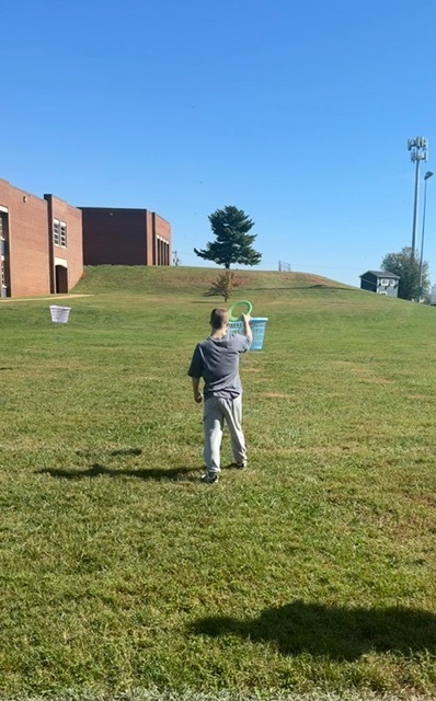 Person holding blue bucket standing on grass, one white bucket in background, building and trees in distance.