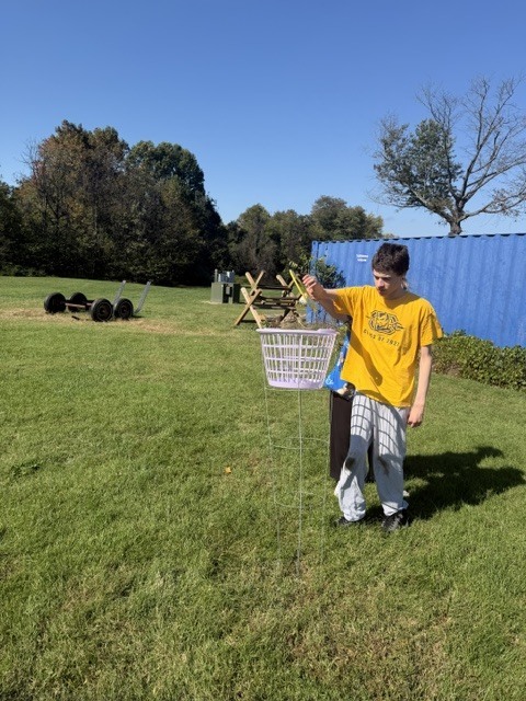 A person in a yellow shirt holds a basket filled with items in a grassy field.