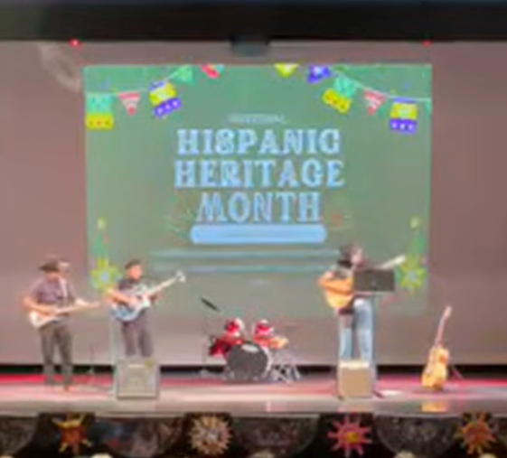 People on stage playing guitars and drums. A large screen behind them reads "HISPANIC HERITAGE MONTH".