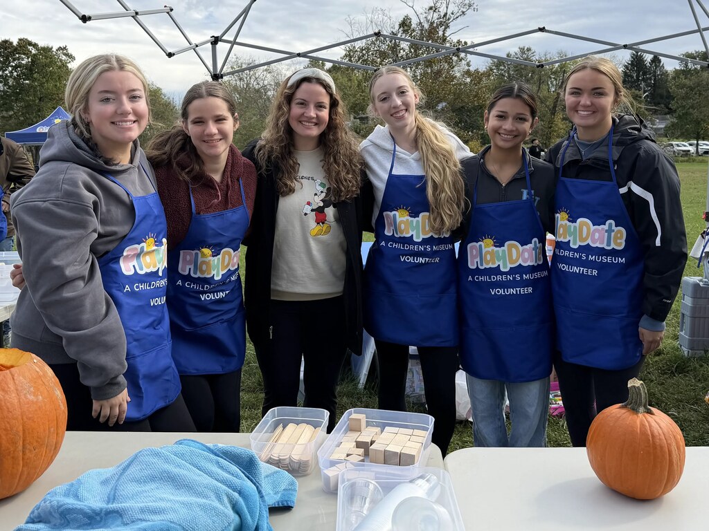 Five people pose under a tent. One holds a pumpkin. All wear aprons. A table has pumpkins, a cloth, and containers.