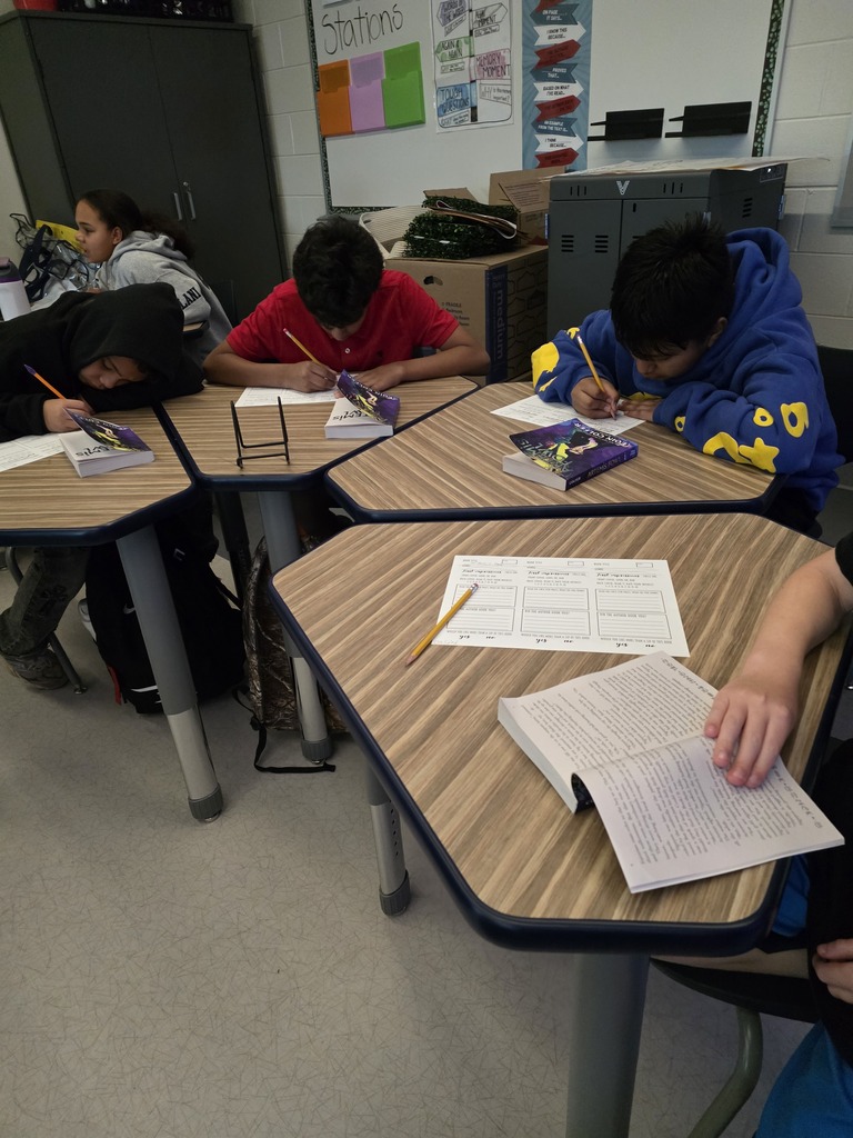 CMS students work independently during a book tasting activity, reading books and completing worksheets at their desks. Several students are writing responses while copies of Artemis Fowl rest on their desks.