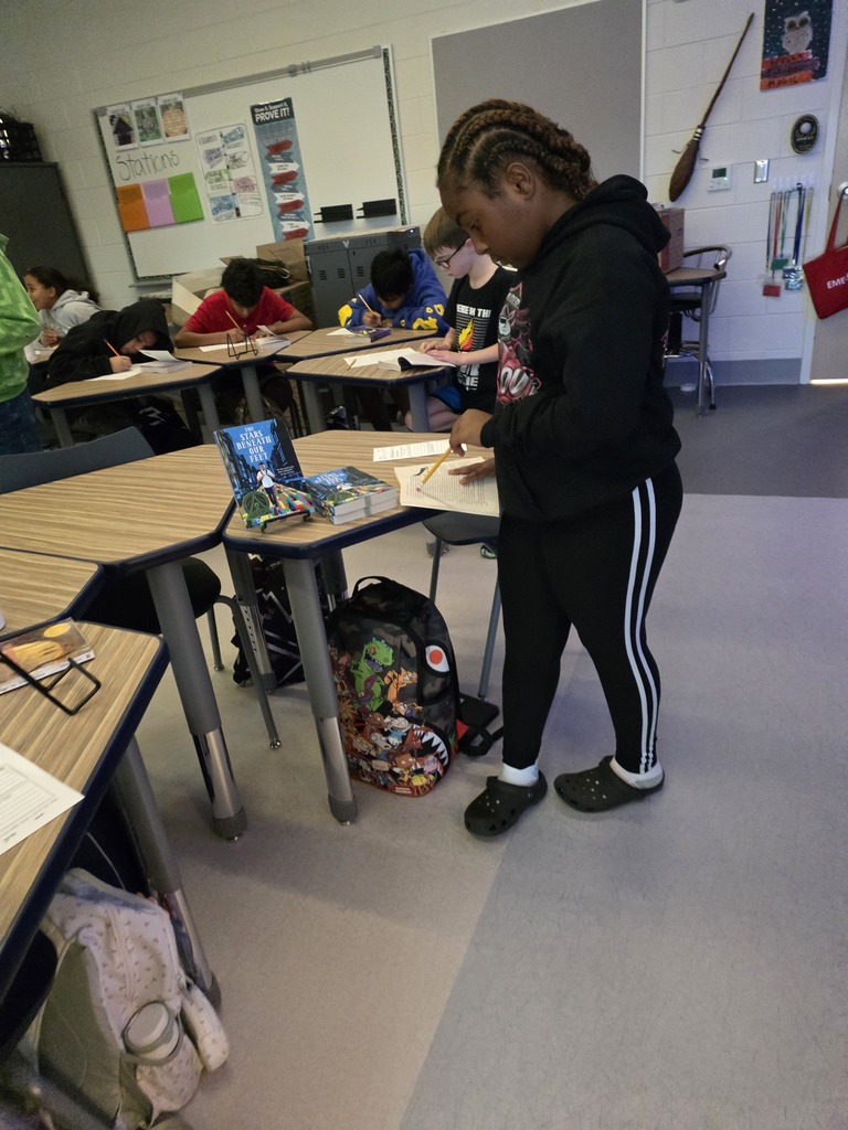 CMS students participate in a book tasting activity, reading and completing worksheets at classroom stations. A student in the foreground examines a book beside a display of The Stars Beneath Our Feet, while classmates work at desks in the background.